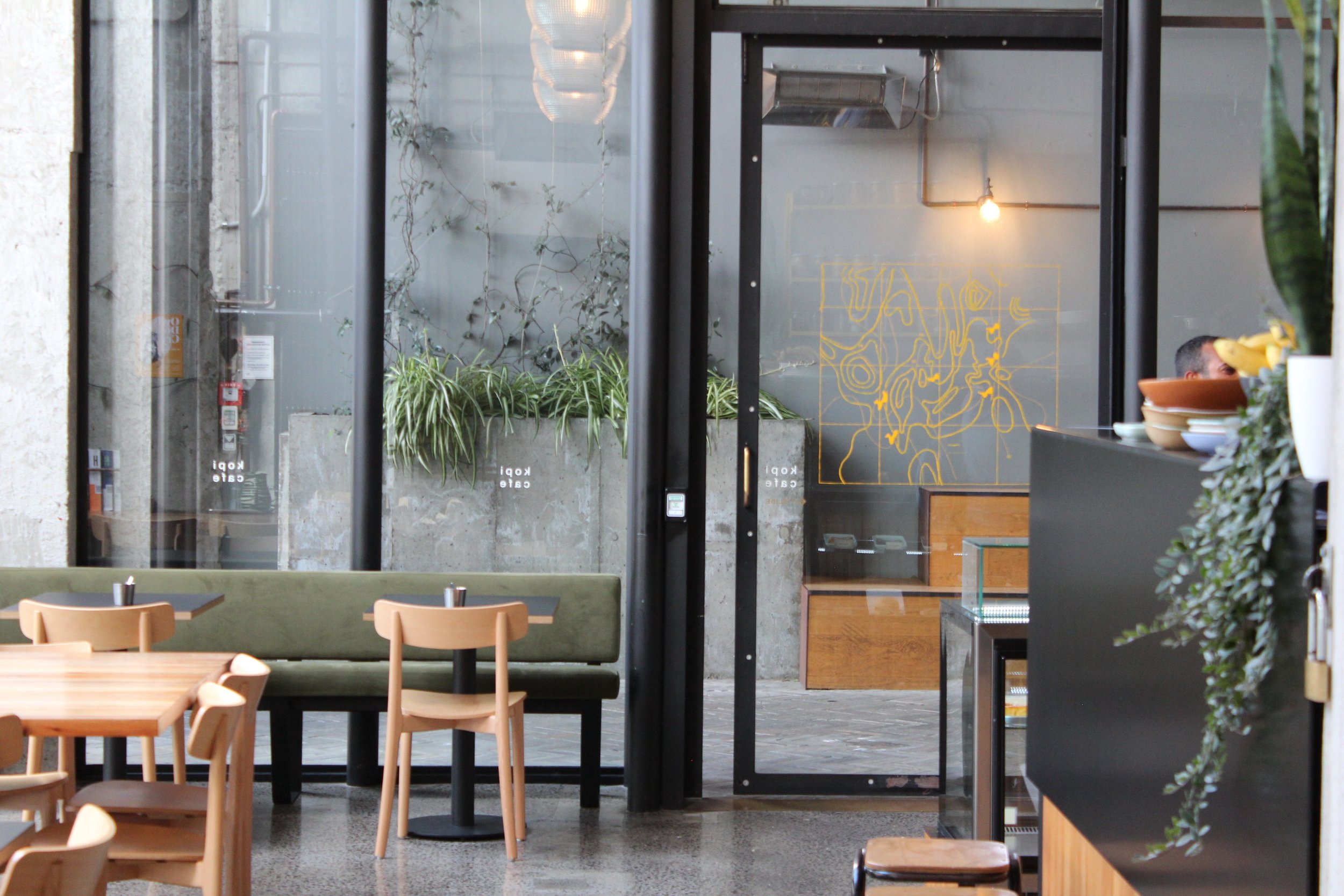 Inside Kopi Cafe with concrete columns, glass walls, timber seating, and yellow shelving.