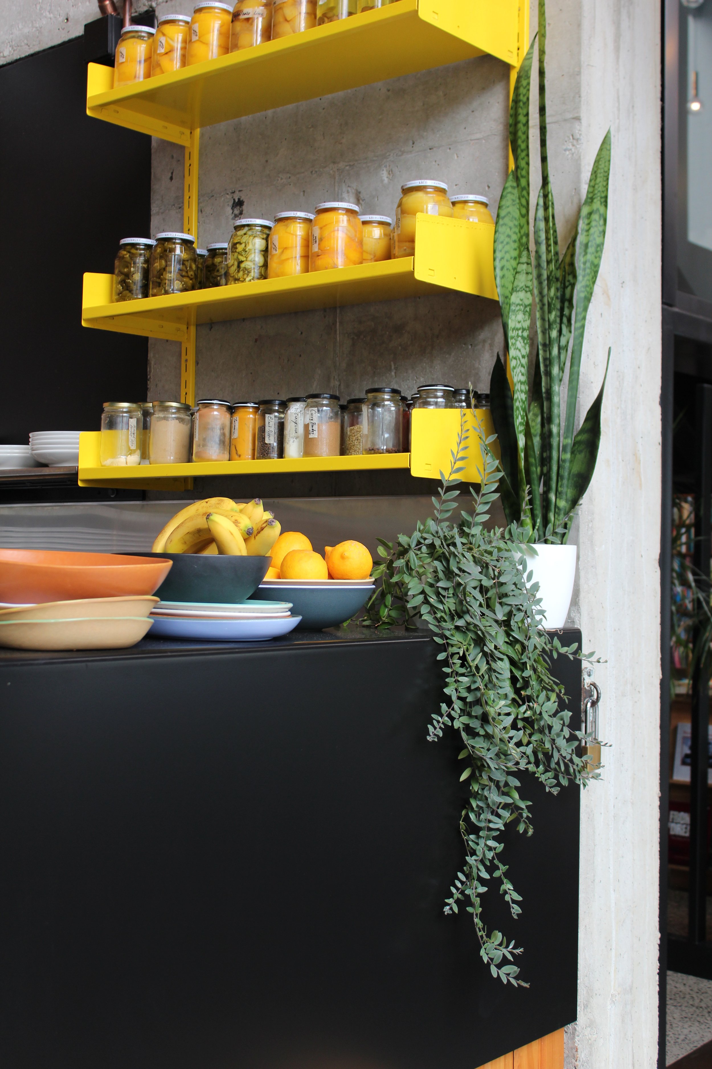 Kopi Cafe shelf detail with yellow shelving, concrete wall, plants, fruit, and black counter.